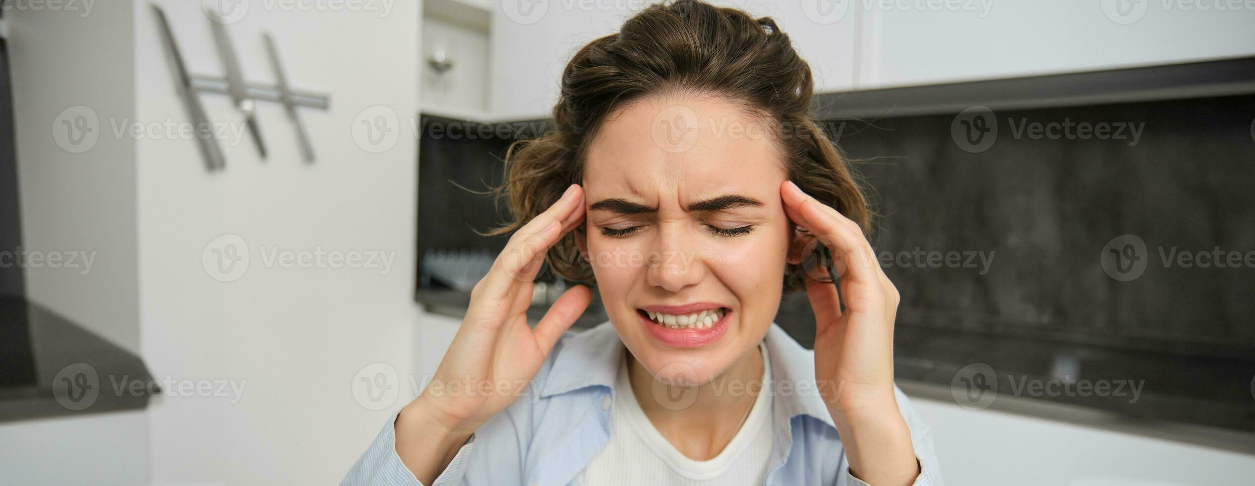 Portrait of woman with headache, sits in kitchen, touches head and grimaces, has painful migraine photo