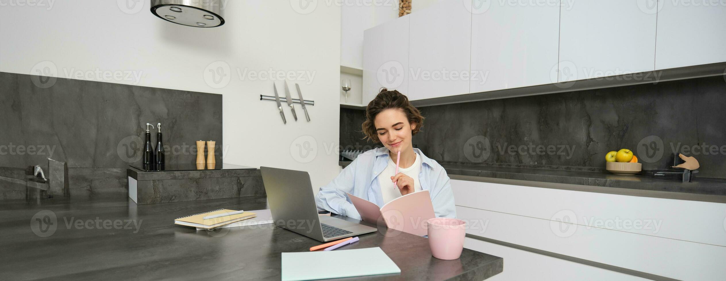 Portrait of young beautiful woman, studies at home, flips her workbook, works from home, uses laptop to learn on remote, makes notes during webinar photo