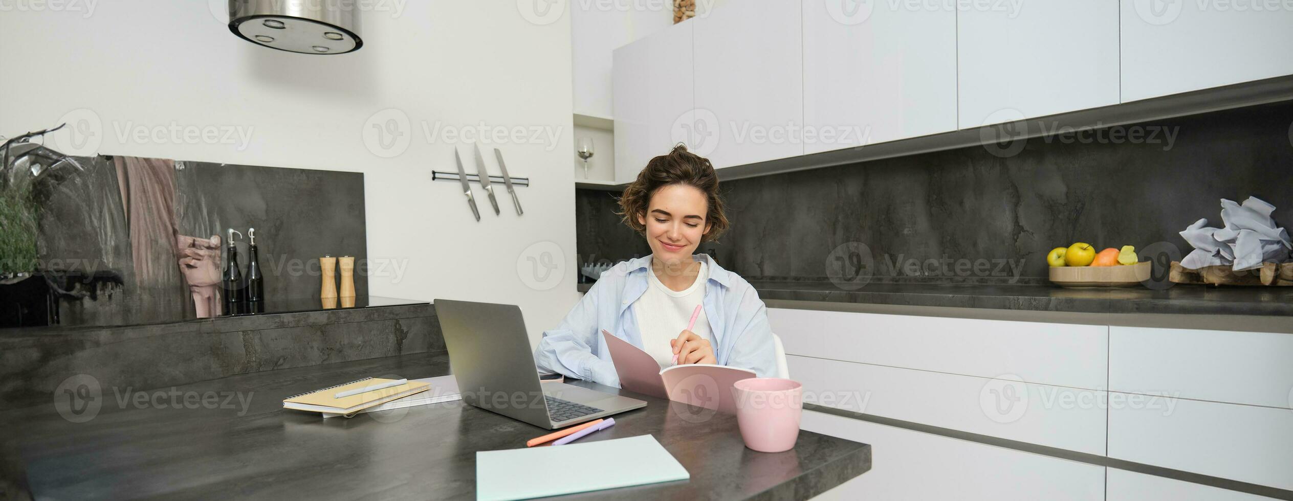 Portrait of brunette woman does homework, works from home, prepares for exam in kitchen, sits with notebook and laptop photo