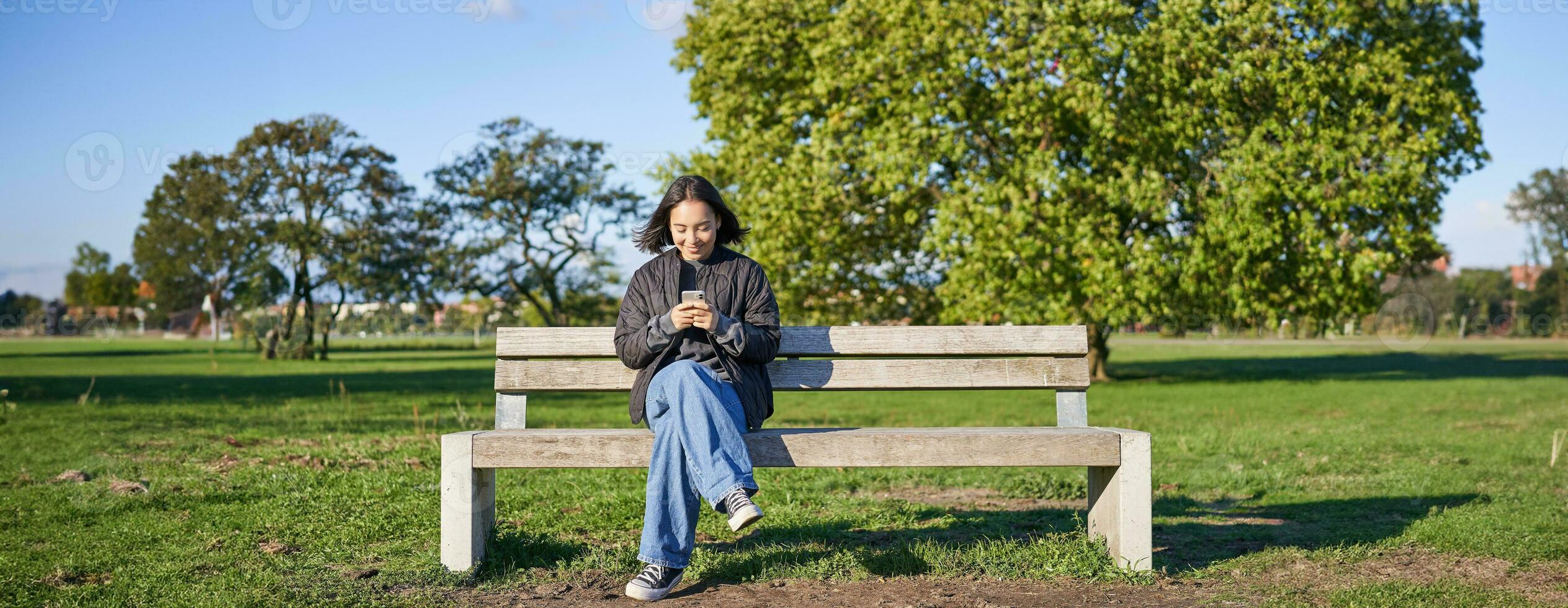 Young woman sitting alone on bench in park, using mobile phone, looking at screen, texting, sending messages in app chat photo