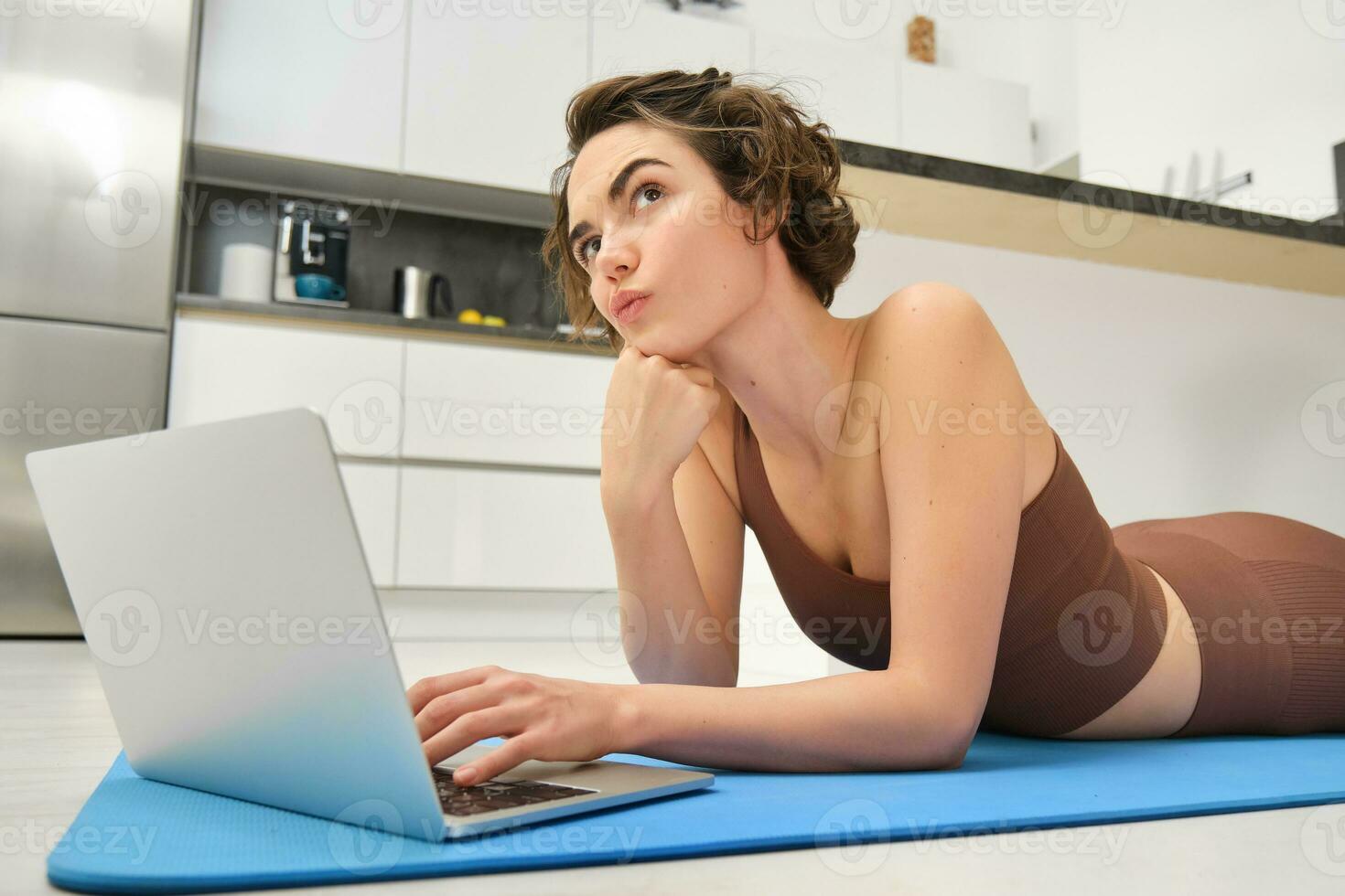 Portrait of young sportswoman, lying on floor at home, using yoga mat, workout, thinking while using laptop, searching for online gym exercises for training indoors photo
