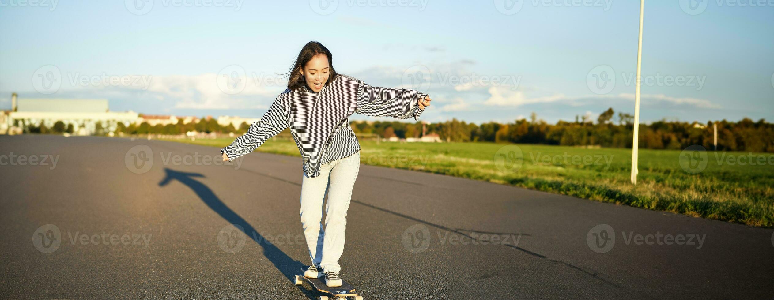 Vertical portrait of happy asian girl enjoying skateboard fun day out. Smiling korean skater on longboard, riding along empty street on sunny day photo