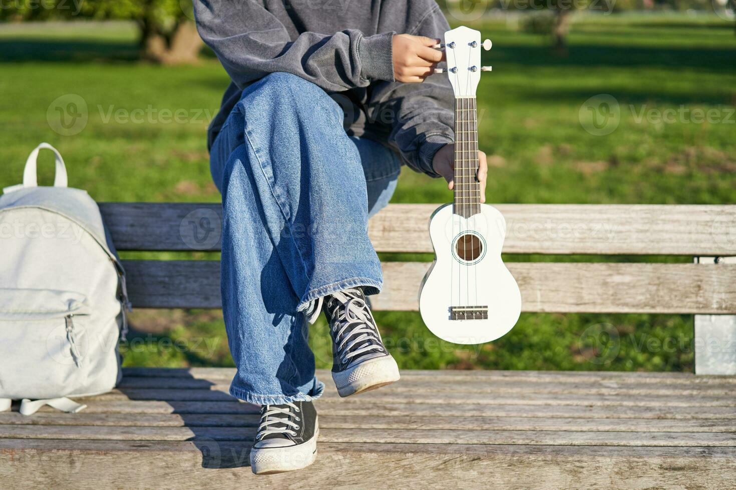 recortado Disparo de joven niña en zapatillas y vaqueros, manos participación ukelele musical instrumento mientras ella se sienta en banco en verde soleado parque foto