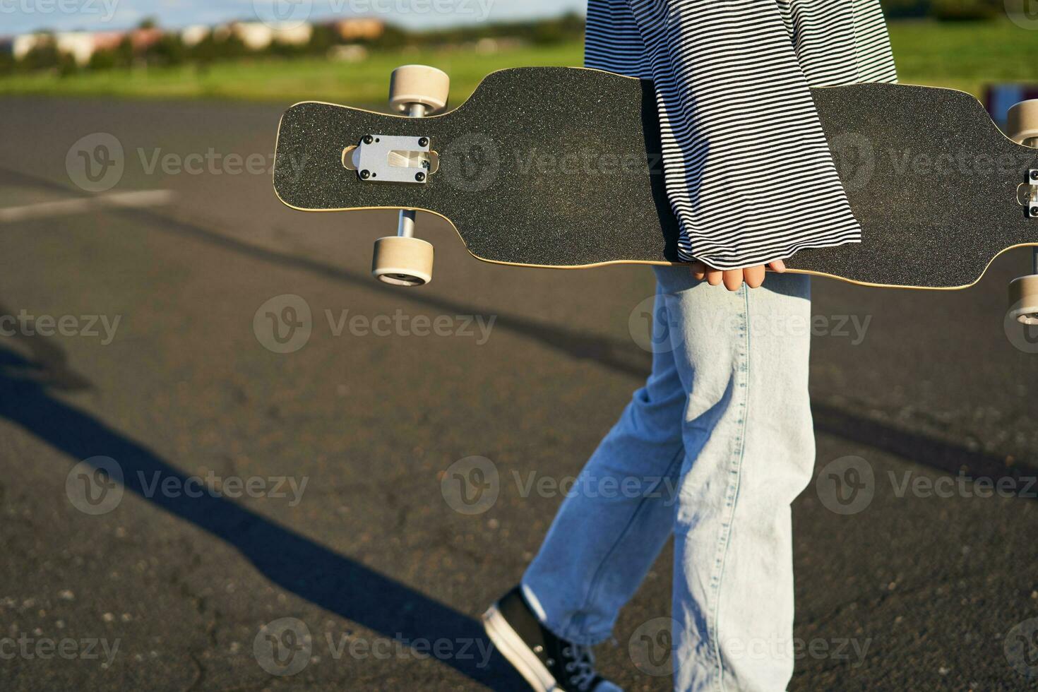 Cropped shot of teen skater girl, hands holding longboard, walking with skateboard on concrete road photo