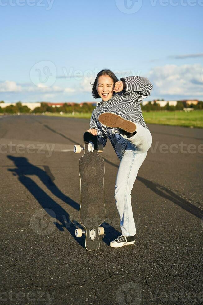 Vertical shot of asian girl feeling excited, skating on longboard, jumping and posing with skateboard, standing with cruiser on empty road, having fun outdoors photo