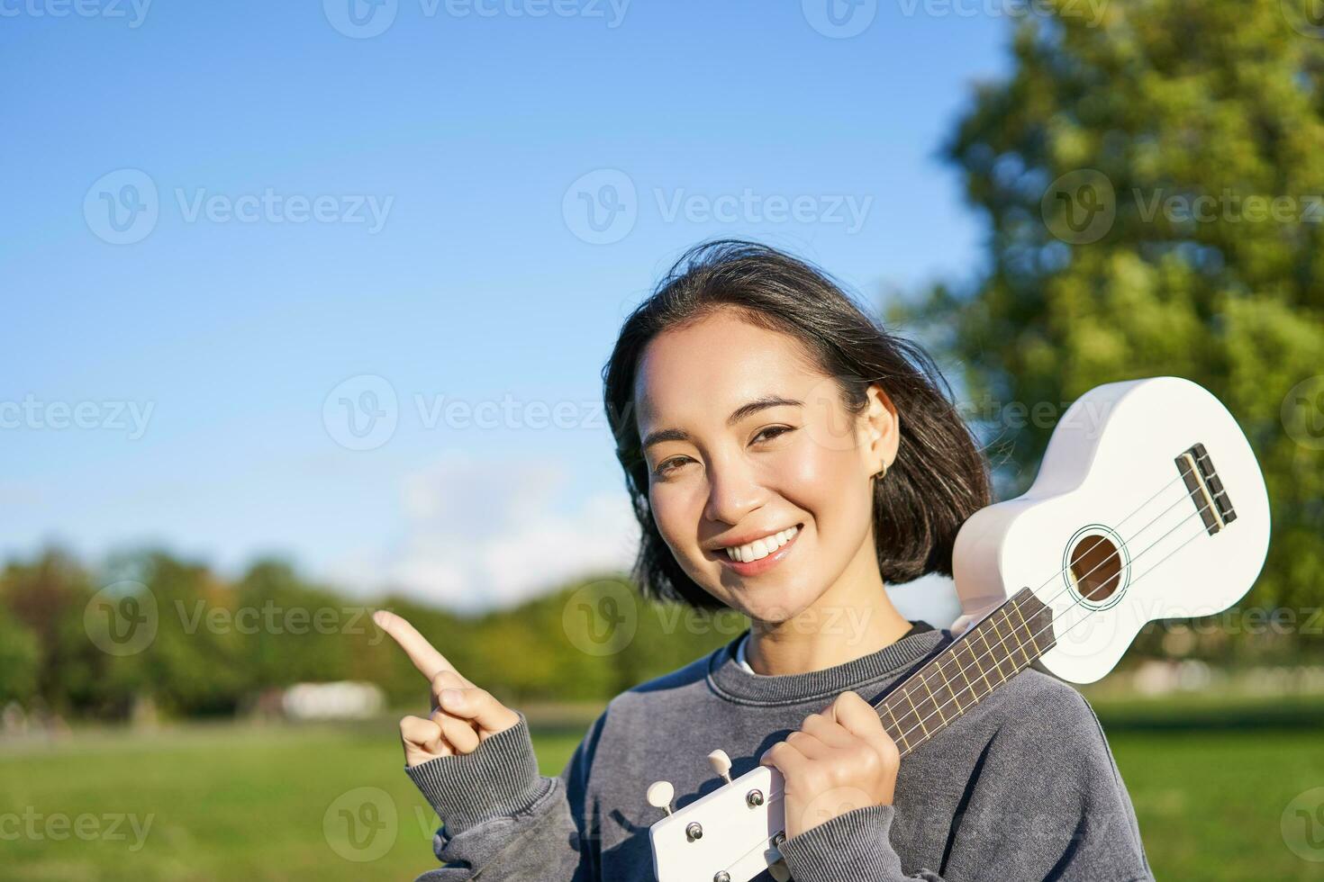 Portrait of asian smiling girl, holding ukulele over shoulder, pointing finger at copy space, banner or logo photo