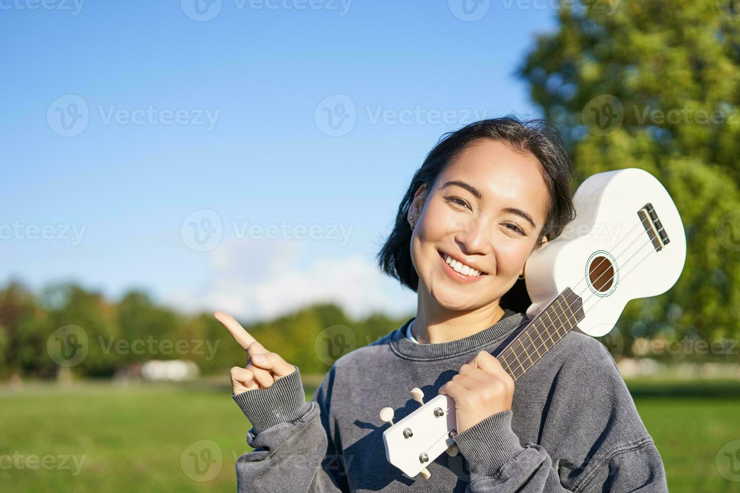 Portrait of asian smiling girl, holding ukulele over shoulder, pointing finger at copy space, banner or logo photo