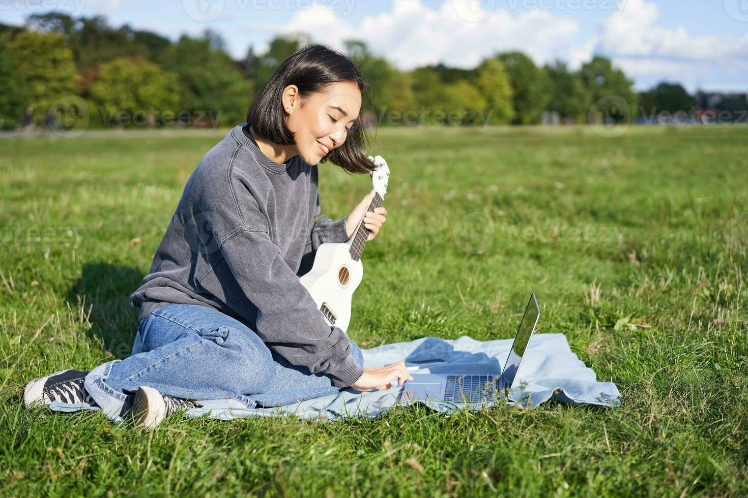 Smiling asian girl learns how to play ukulele via laptop, online video tutorials, sitting on grass in park with musical instrument photo