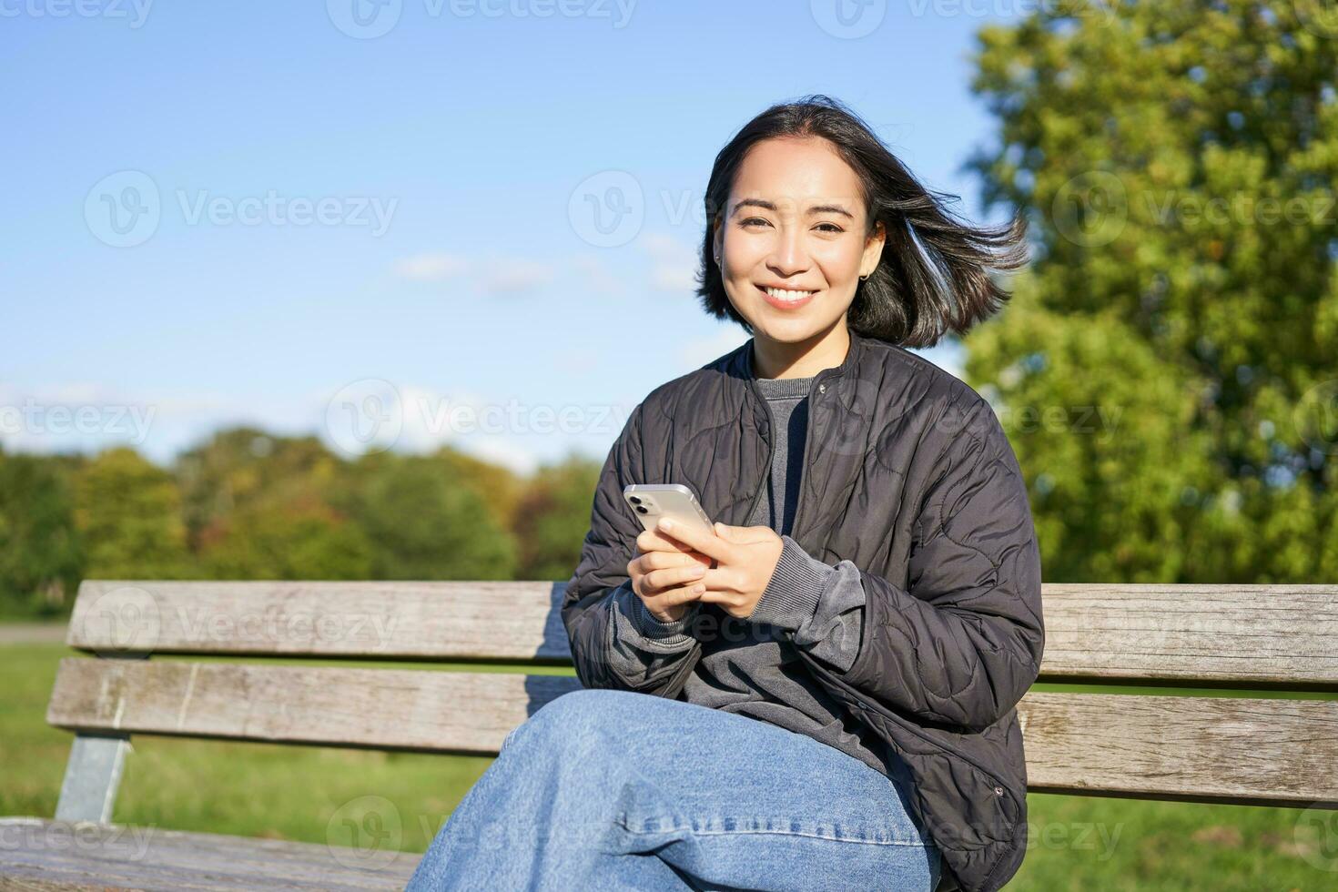 Technology and people. Young woman using smartphone, sitting outdoors on bench in park, looking at smartphone, sending message or networking photo