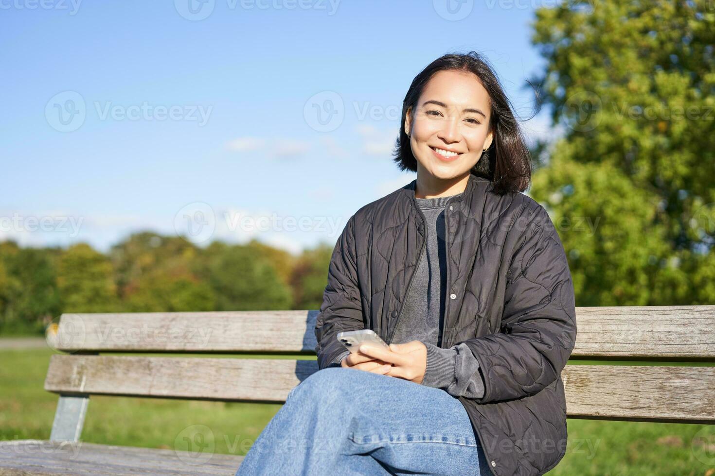 Technology and people. Young woman using smartphone, sitting outdoors on bench in park, looking at smartphone, sending message or networking photo