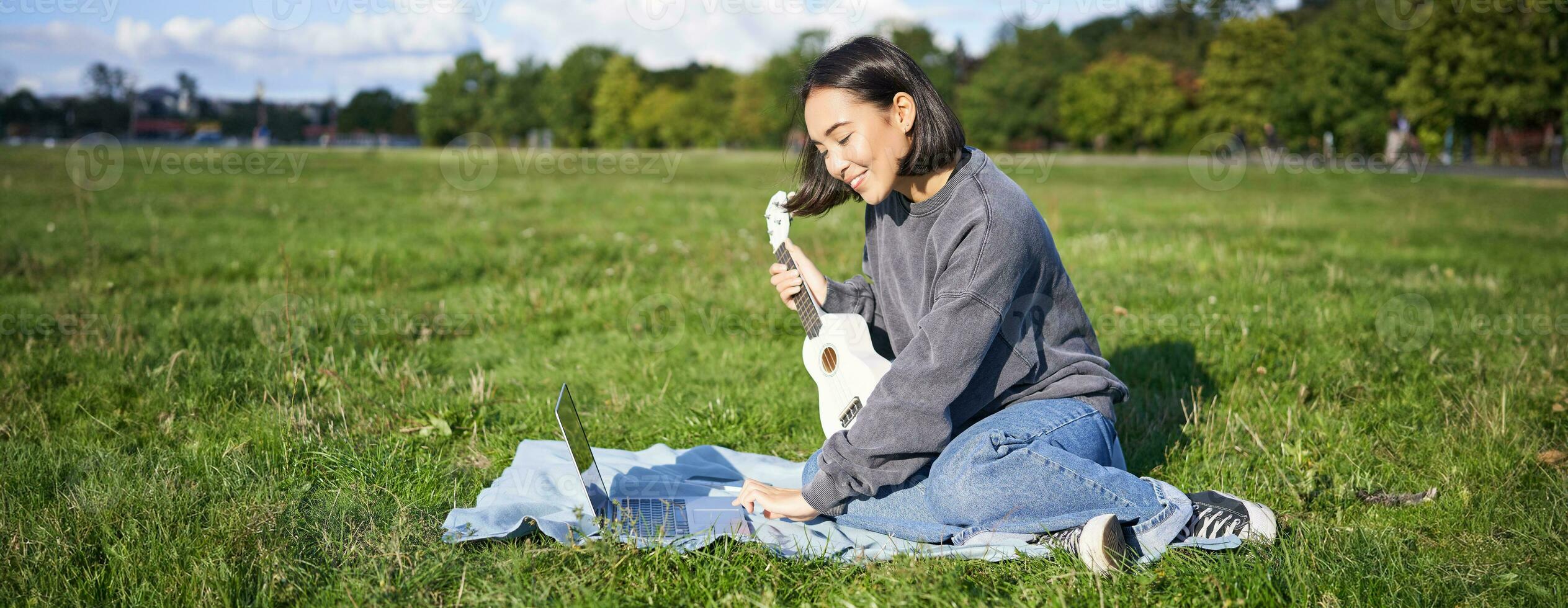 Smiling asian girl learns how to play ukulele via laptop, online video tutorials, sitting on grass in park with musical instrument photo