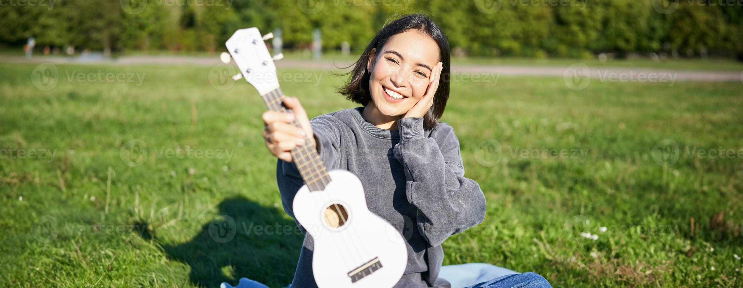 hermosa asiático niña con contento sonrisa, muestra su ukelele, se sienta fuera de en parque en césped, relaja con música foto