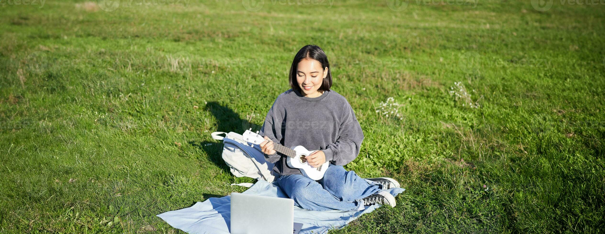 Cute asian girl, musician studying, looks at tutorial on her laptop, online tutor shows how to play ukulele, student sits on grass in park on sunny day photo