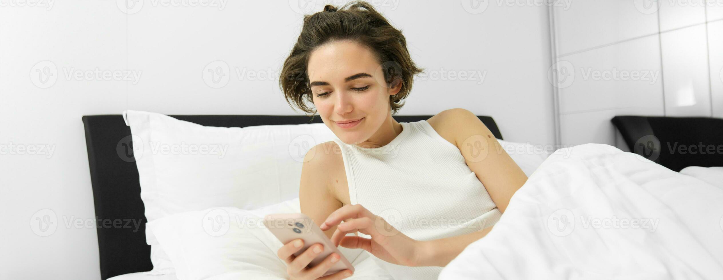 Image of female model in bed, looking at smartphone, sending a message, turns on alarm clock on mobile phone, goes to sleep in her bedroom photo