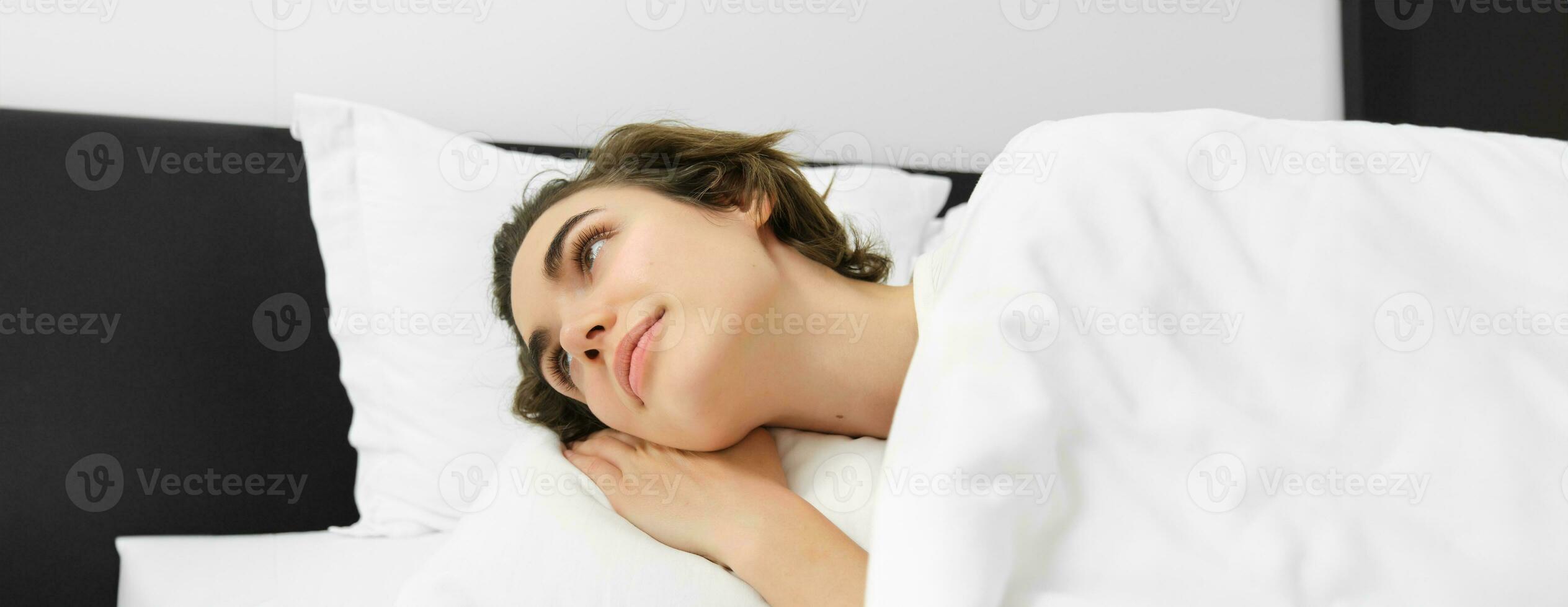 Close up portrait of smiling young woman, lying on white pillow, covered in linen sheets, looking aside at night table, waking up in hotel suit photo