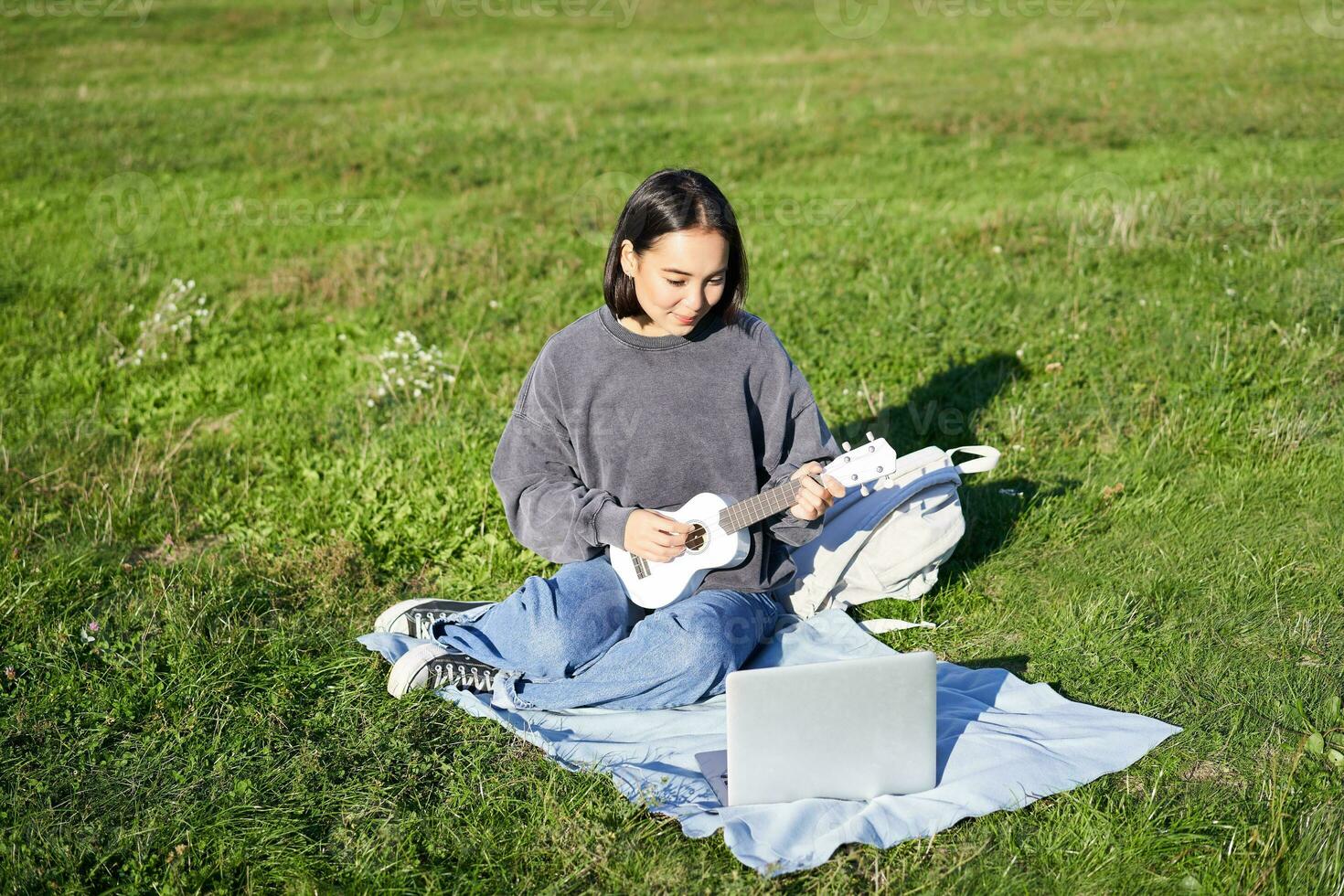 Cute asian girl, musician studying, looks at tutorial on her laptop, online tutor shows how to play ukulele, student sits on grass in park on sunny day photo