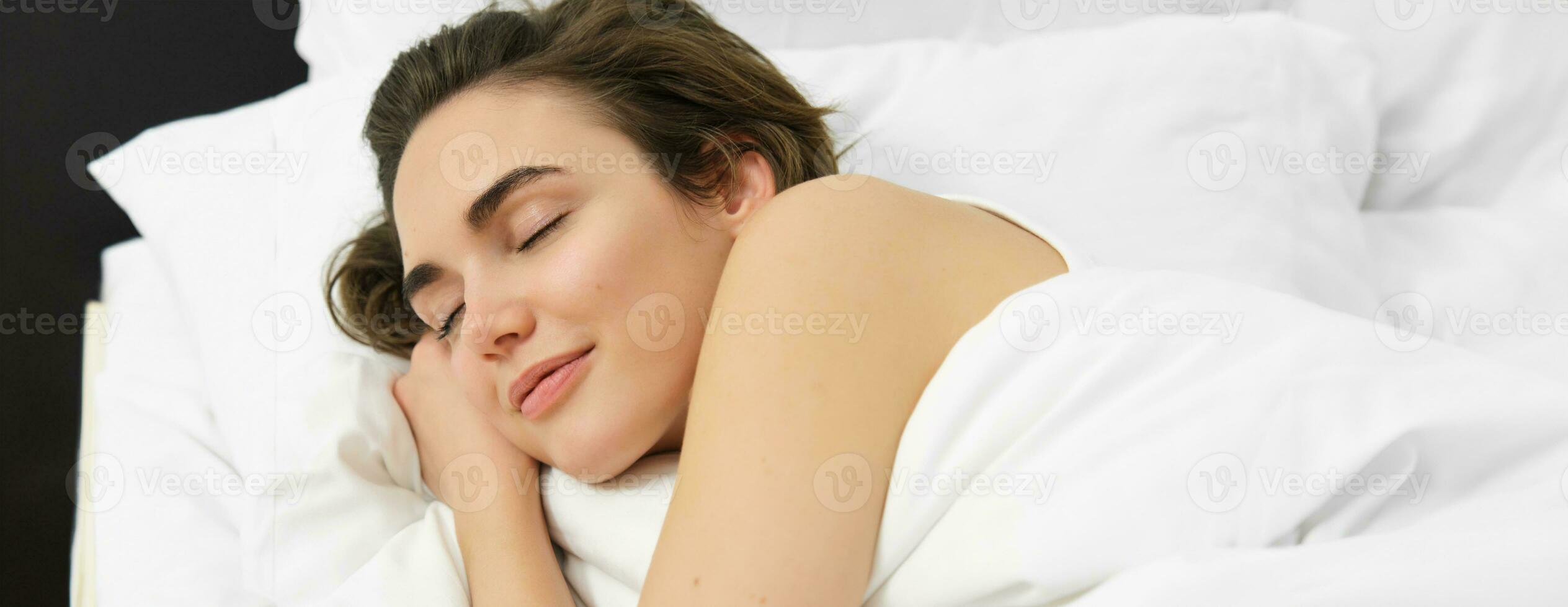 Close up portrait of young woman sleeping in her bed on soft white pillow, eyes closed, smiling white dreaming photo