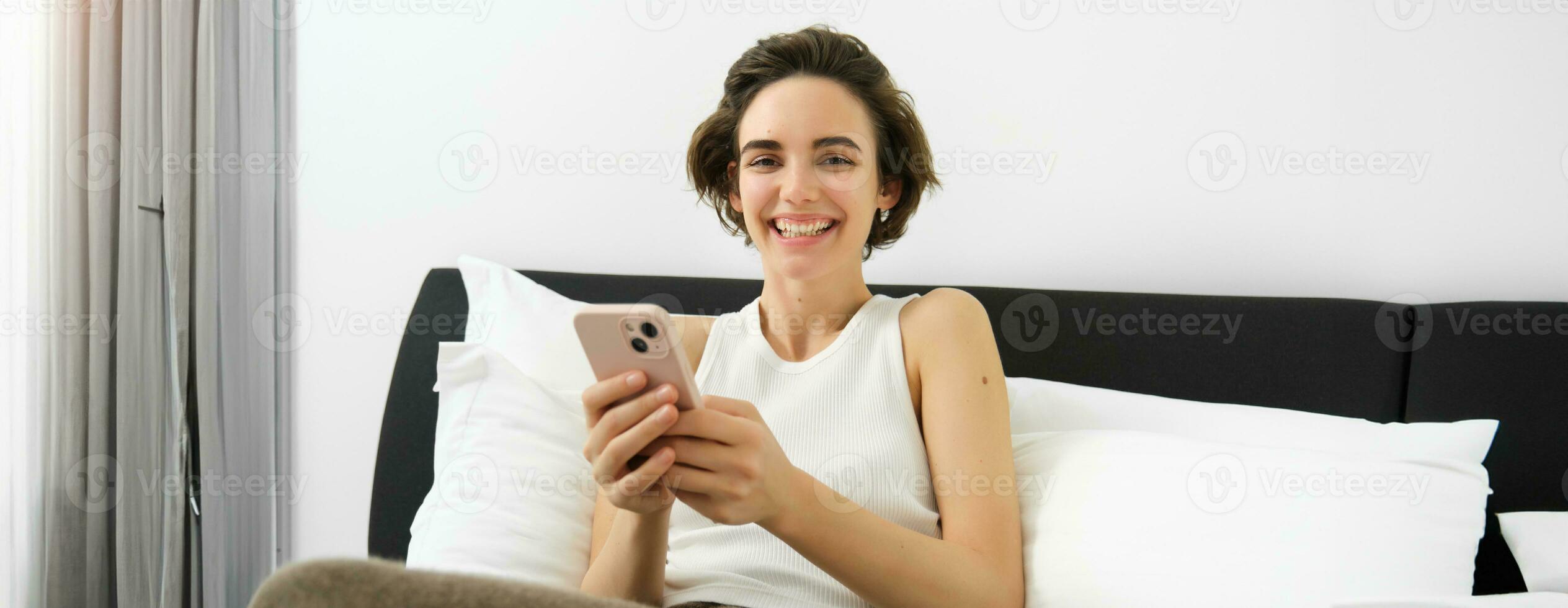 Portrait of smiling brunette woman resting in her bedroom, using mobile phone and lying in bed in her home outfit, messaging, sending message on smartphone app photo
