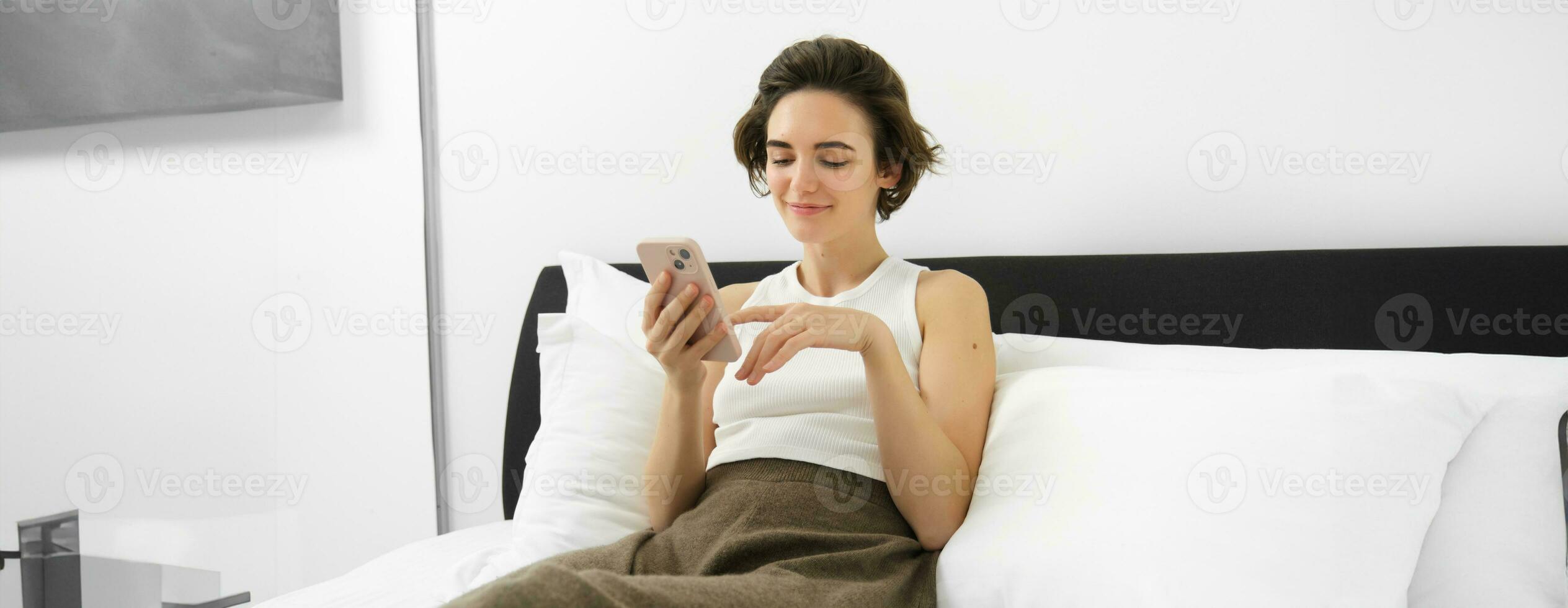 Portrait of cute female model in bedroom, holding phone, typing message on smartphone, resting in bed and smiling photo