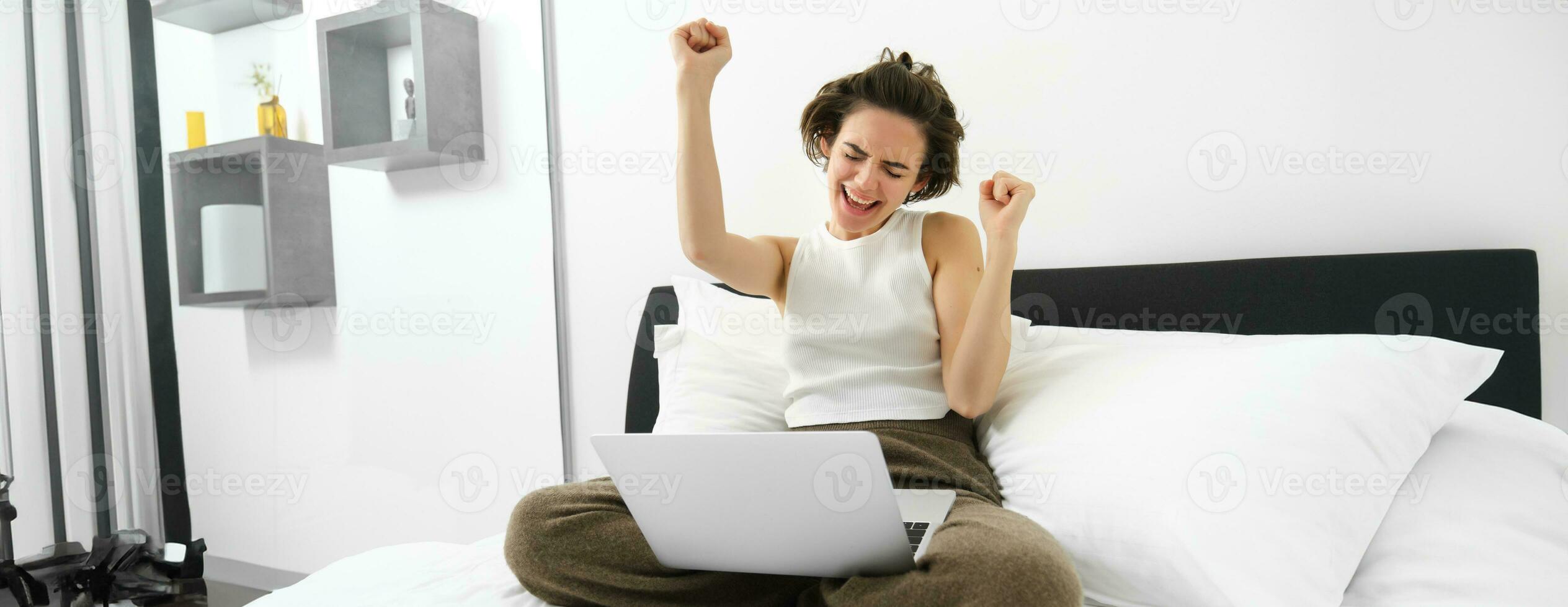 retrato de contento mujer alegría, sentado con computadora y haciendo ganador bailar, tiene ordenador portátil en su vueltas, levantamiento manos arriba y celebrando, triunfando con alegría foto