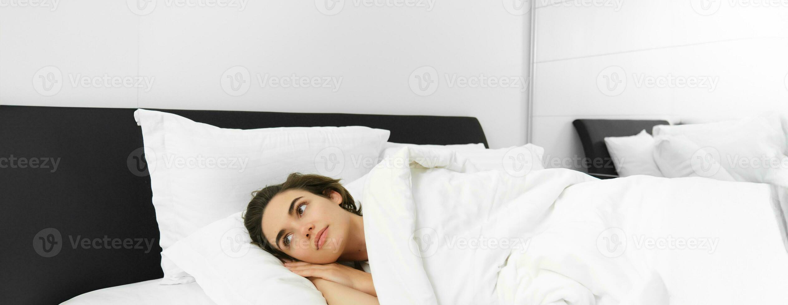 Portrait of dreamy young woman, getting ready for night sleep, lying in bed, covered in white linen sheets, looking aside, waking up in morning photo