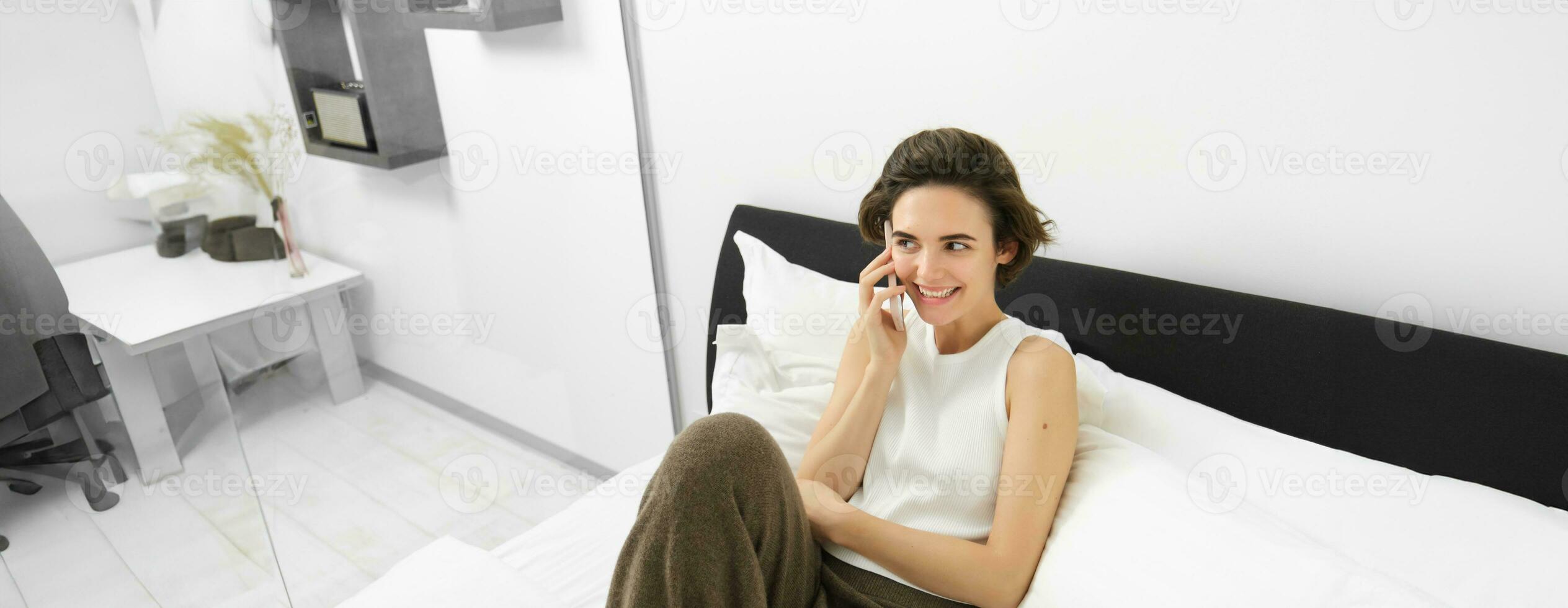 Chatty young woman laughing, talking on phone, calling friend, lying in bed and having conversation with someone, resting in bedroom photo