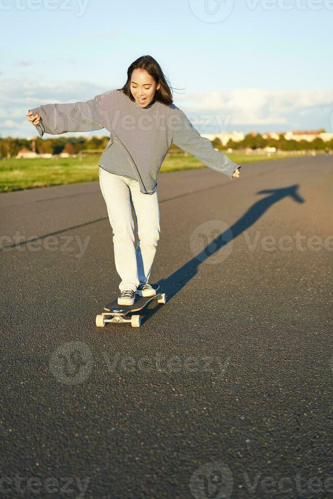 Vertical portrait of happy asian girl enjoying skateboard fun day out. Smiling korean skater on longboard, riding along empty street on sunny day photo