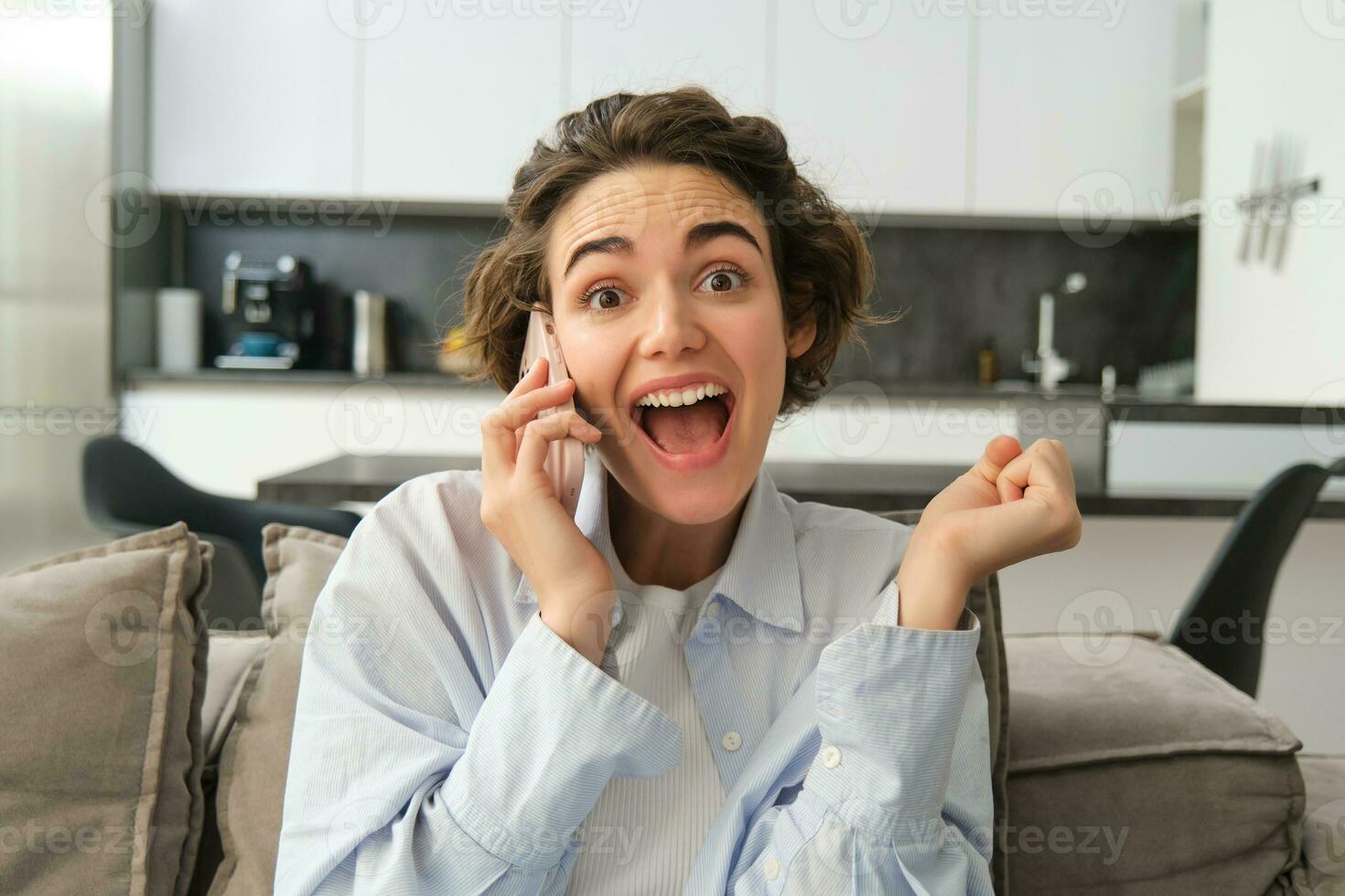 Close up portrait of amazed brunette woman, jumps on sofa from excitement, answers phone call, hear great news over the telephone, reacts surprised and happy photo