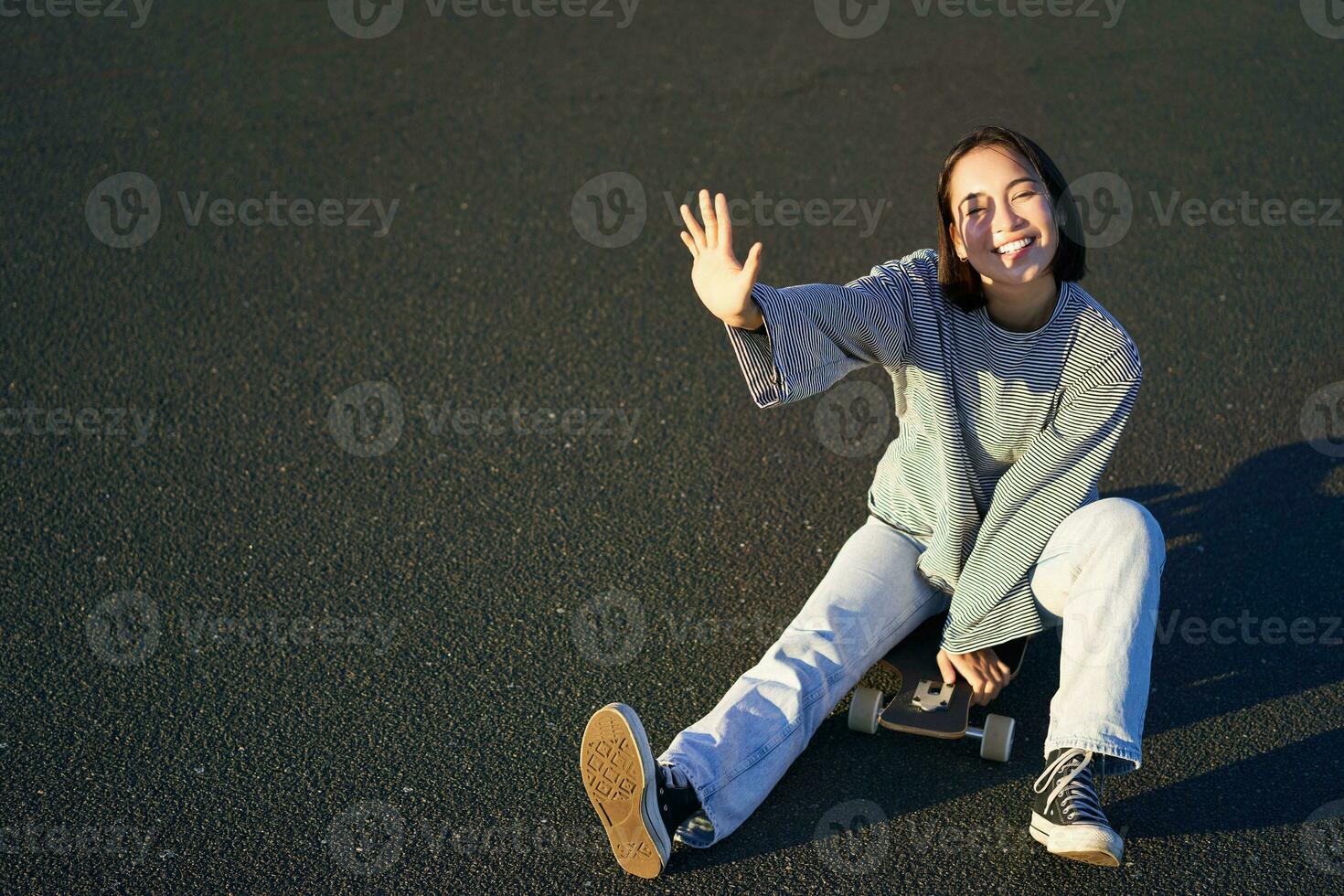 Positive korean girl covers her face from sunlight, sits on skateboard and smiles happily photo