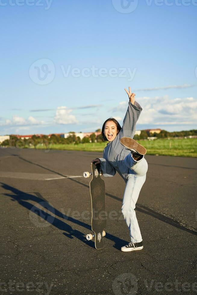 Vertical portrait of smiling asian woman standing on road with longboard, skateboarding on long cruiser, posing on empty road on sunny day photo
