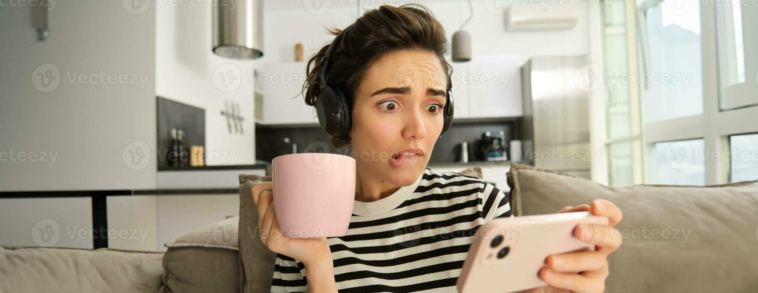 retrato de mujer con preocupado cara expresión, acecho intenso escena en televisión espectáculo en móvil teléfono aplicación, Bebiendo té, ceñudo y curioso a teléfono inteligente, sentado en sofá en vivo habitación foto