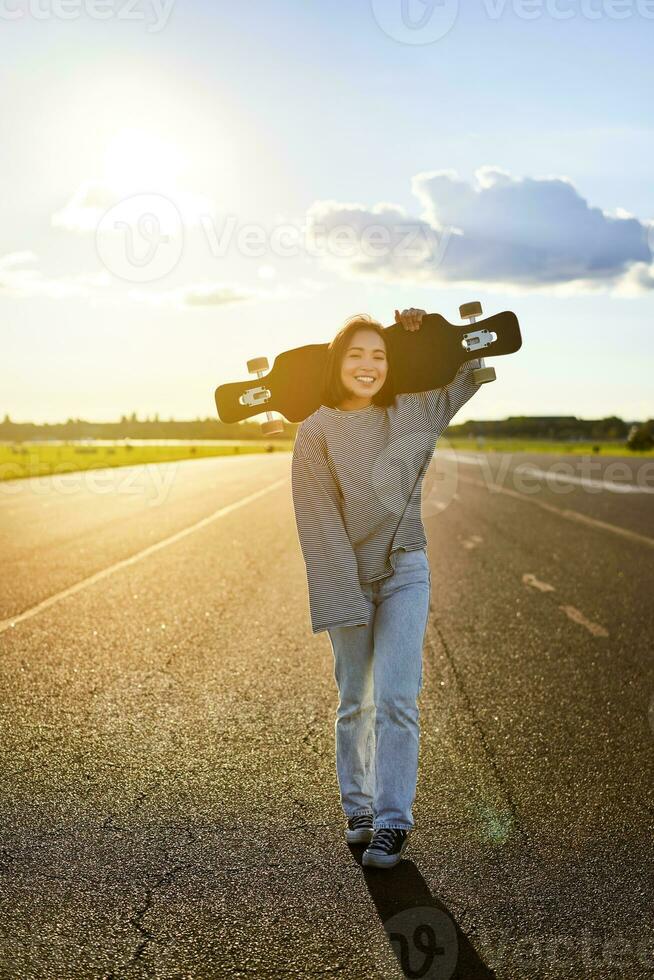 Young asian woman standing with longboard on sunny road, skating in skate park on her cruiser photo