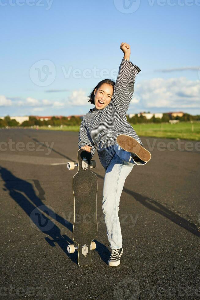 Vertical shot of asian girl feeling excited, skating on longboard, jumping and posing with skateboard, standing with cruiser on empty road, having fun outdoors photo