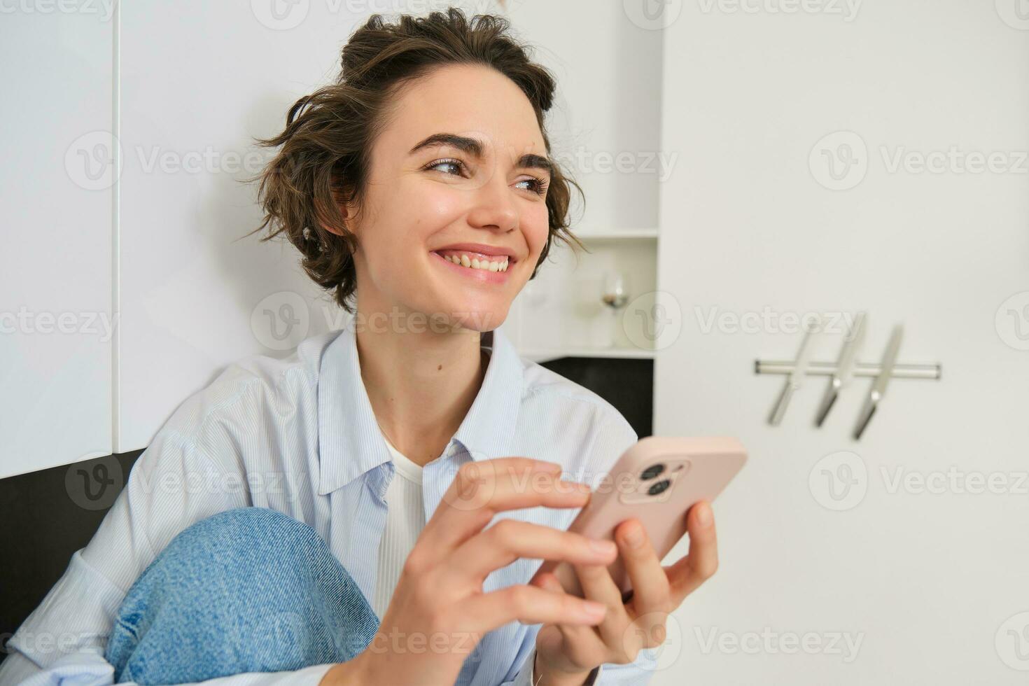 Portrait of smiling beautiful woman, sitting in kitchen with smartphone, looking happy at camera, spending time at home, using mobile phone app photo