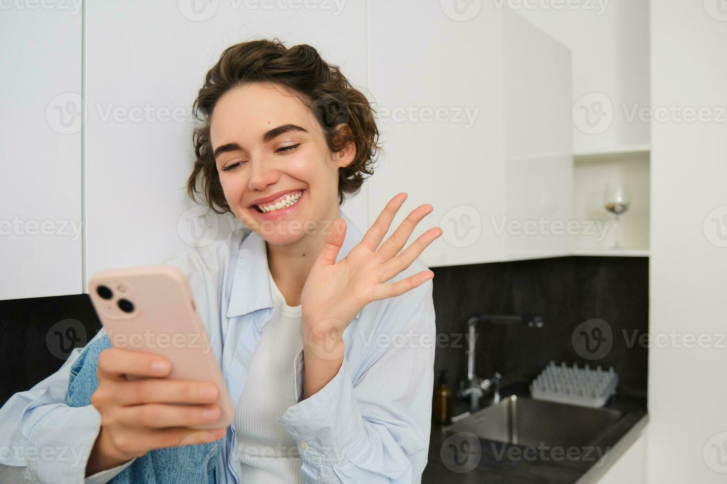 retrato de simpático sonriente morena mujer, sentado a hogar en cocina, mirando a su teléfono inteligente cámara, vídeo chats y olas a móvil teléfono, diciendo Hola a alguien foto