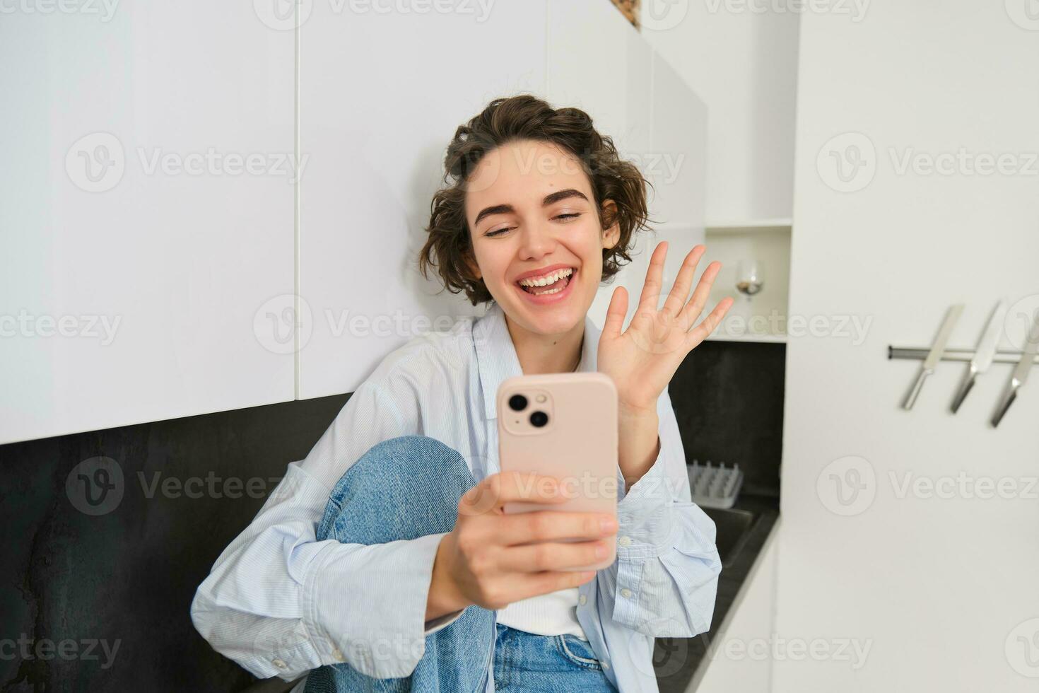 retrato de simpático sonriente morena mujer, sentado a hogar en cocina, mirando a su teléfono inteligente cámara, vídeo chats y olas a móvil teléfono, diciendo Hola a alguien foto
