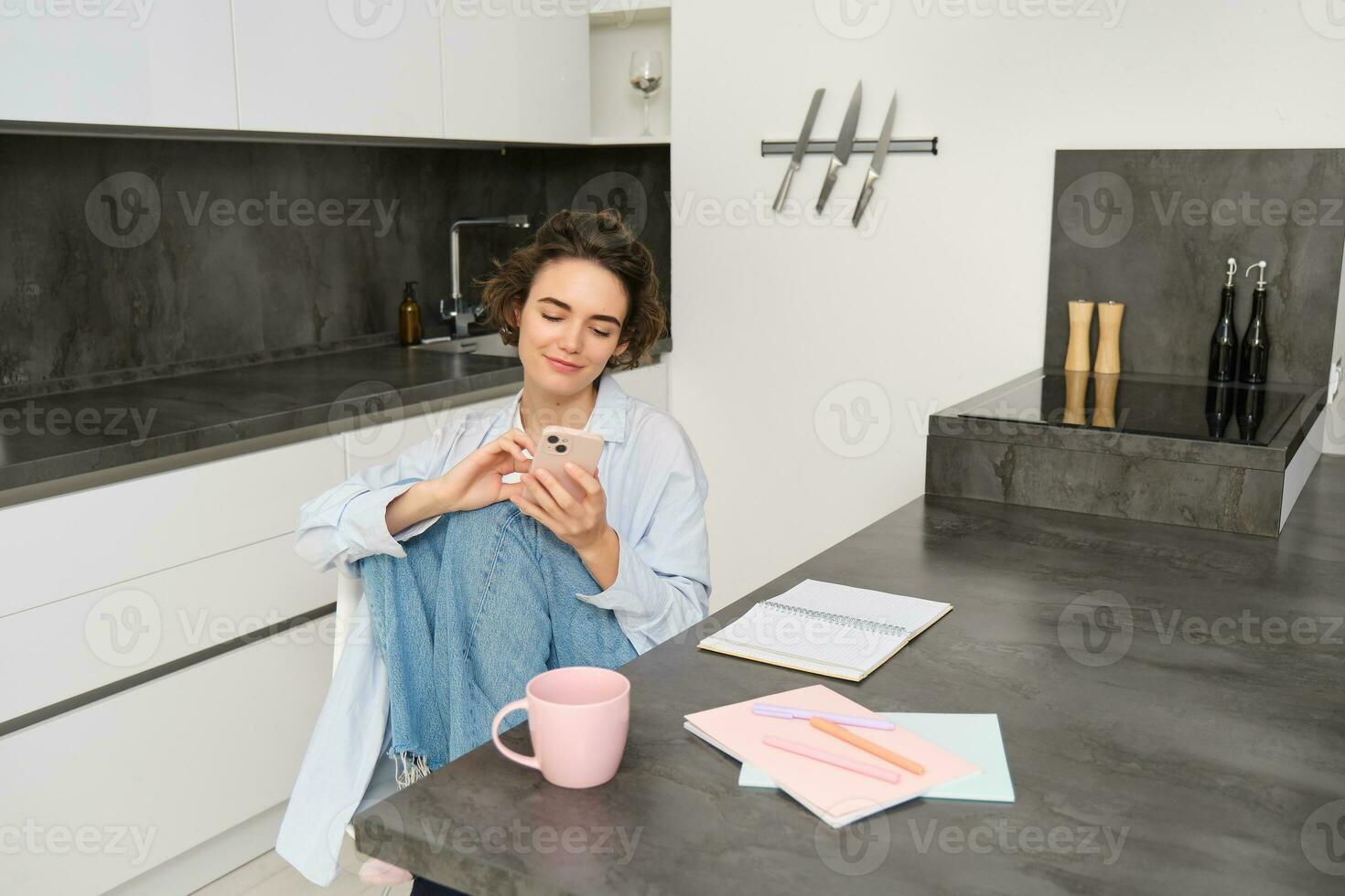Beautiful girl with smartphone, sits at home, does homework, watches smth on mobile phone app photo