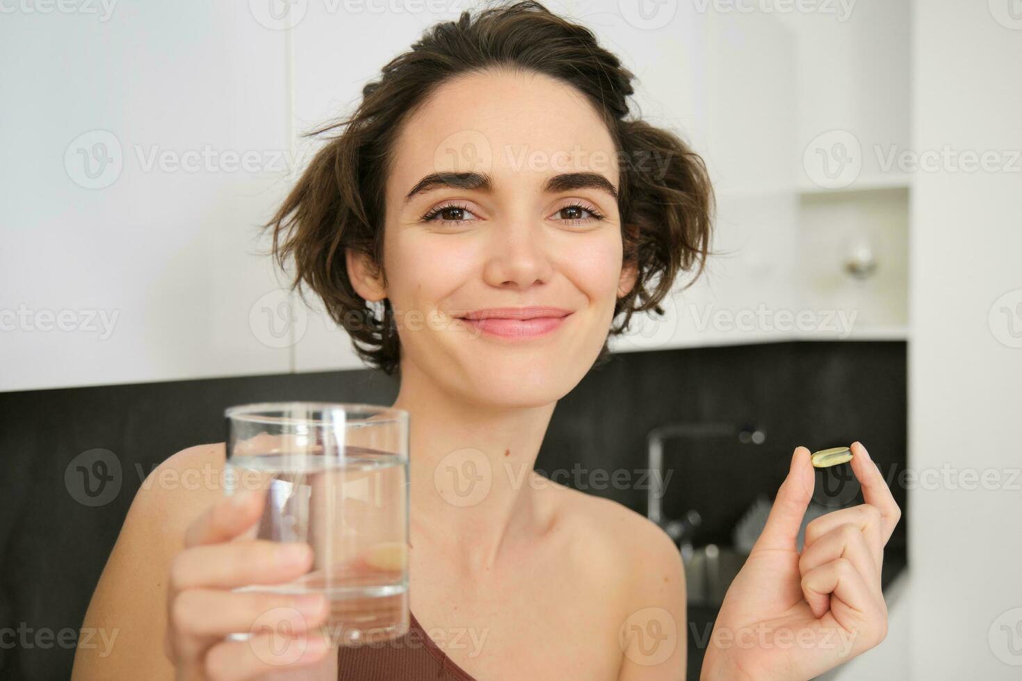 Dietry supplements and healthy lifestyle. Young woman taking vitamin C, D omega-3 with glass of water, standing in activewear, drinking after workout training in her kitchen photo