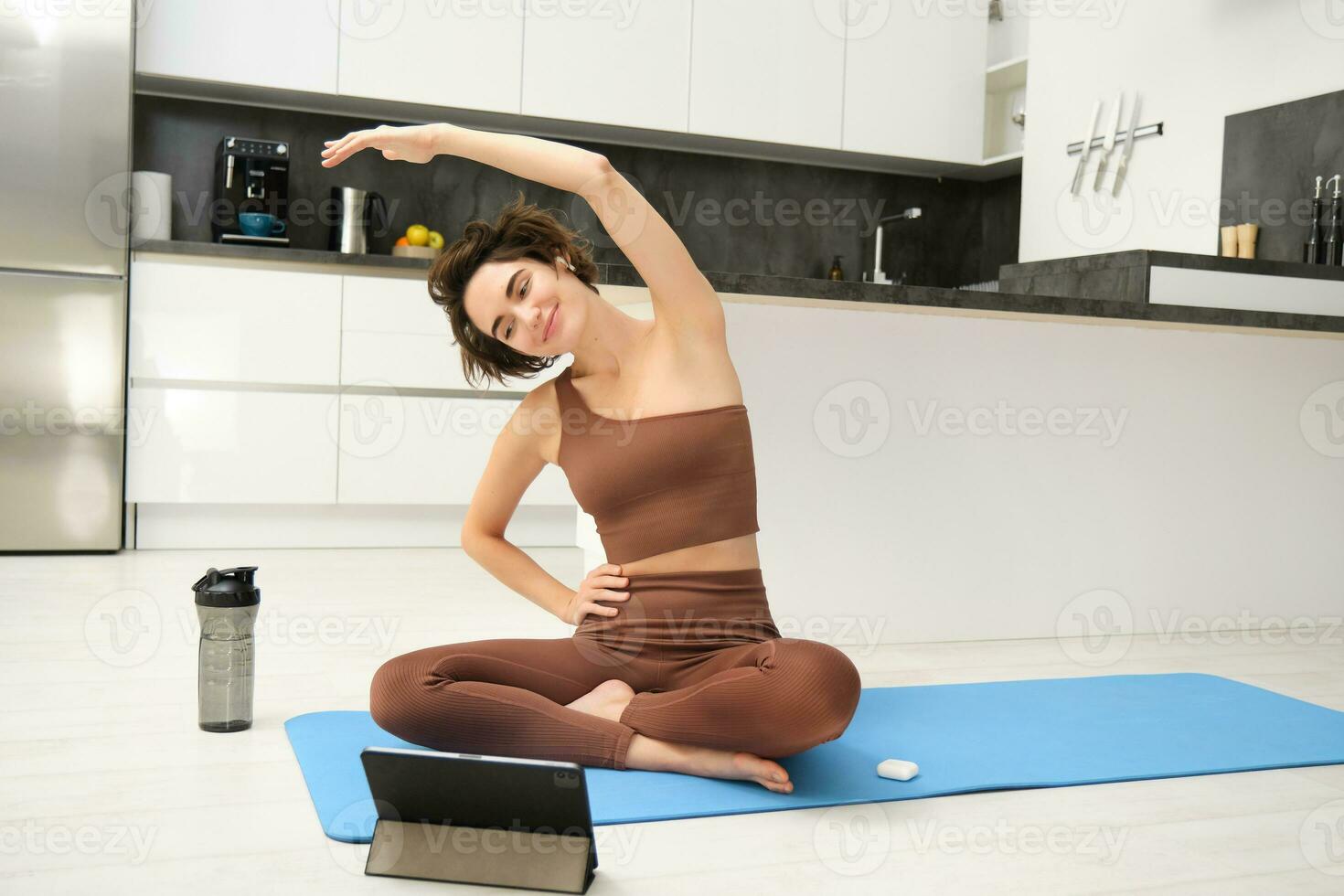 Active young woman, vlogger does sports, records her workout training from  home on smartphone camera, posing for selfie inside her house, sits on  rubber yoga mat in blue leggings and sportsbra 35338950