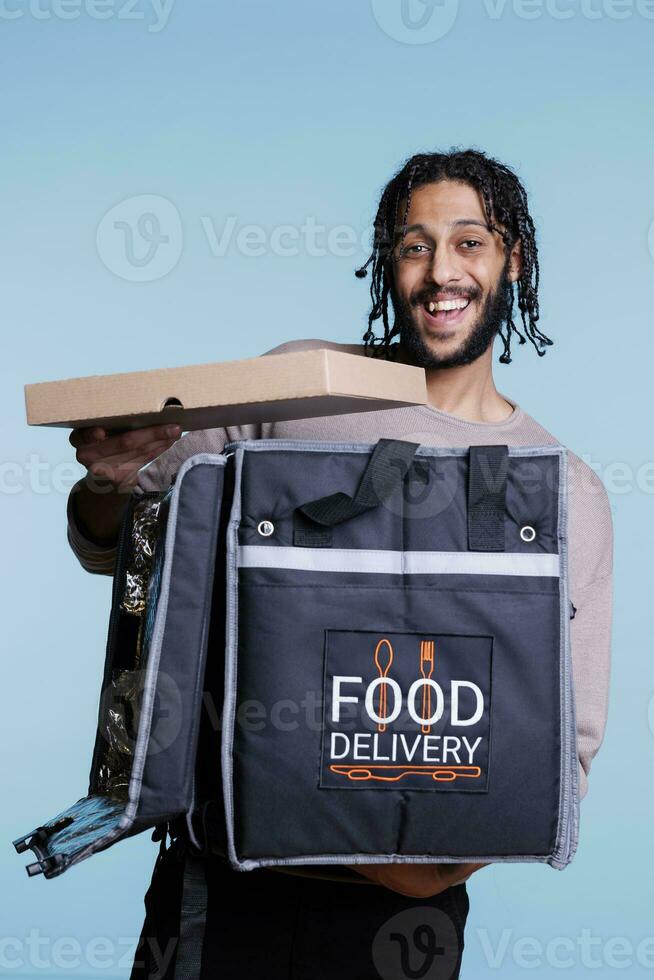 contento árabe mensajero participación Pizza caja, entregando rápido comida y mirando a cámara con alegre expresión. despreocupado pizzería restaurante repartidor en pie con térmico mochila retrato foto