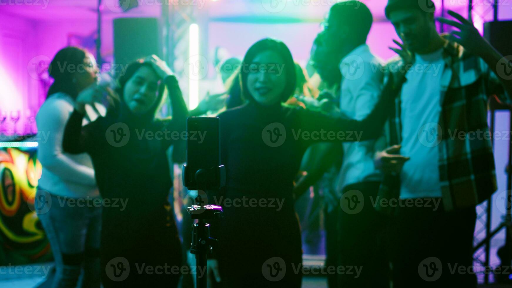 Woman filming vlog for social media, using smartphone at nightclub on dance floor. Young adult recording fun video of friends dancing and partying together, entertainment. Handheld shot. photo