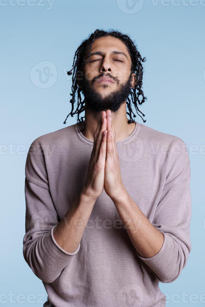 Arab man praying to god with folded hands and making wish with closed eyes. Young person with hopeful facial expression of peace and spirituality pleading for forgiveness concept photo