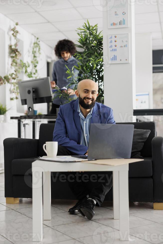 Smiling successful arab businessman working on laptop in coworking space portrait. Happy confident unicorn business entrepreneur sitting on couch at office workplace and looking at camera photo