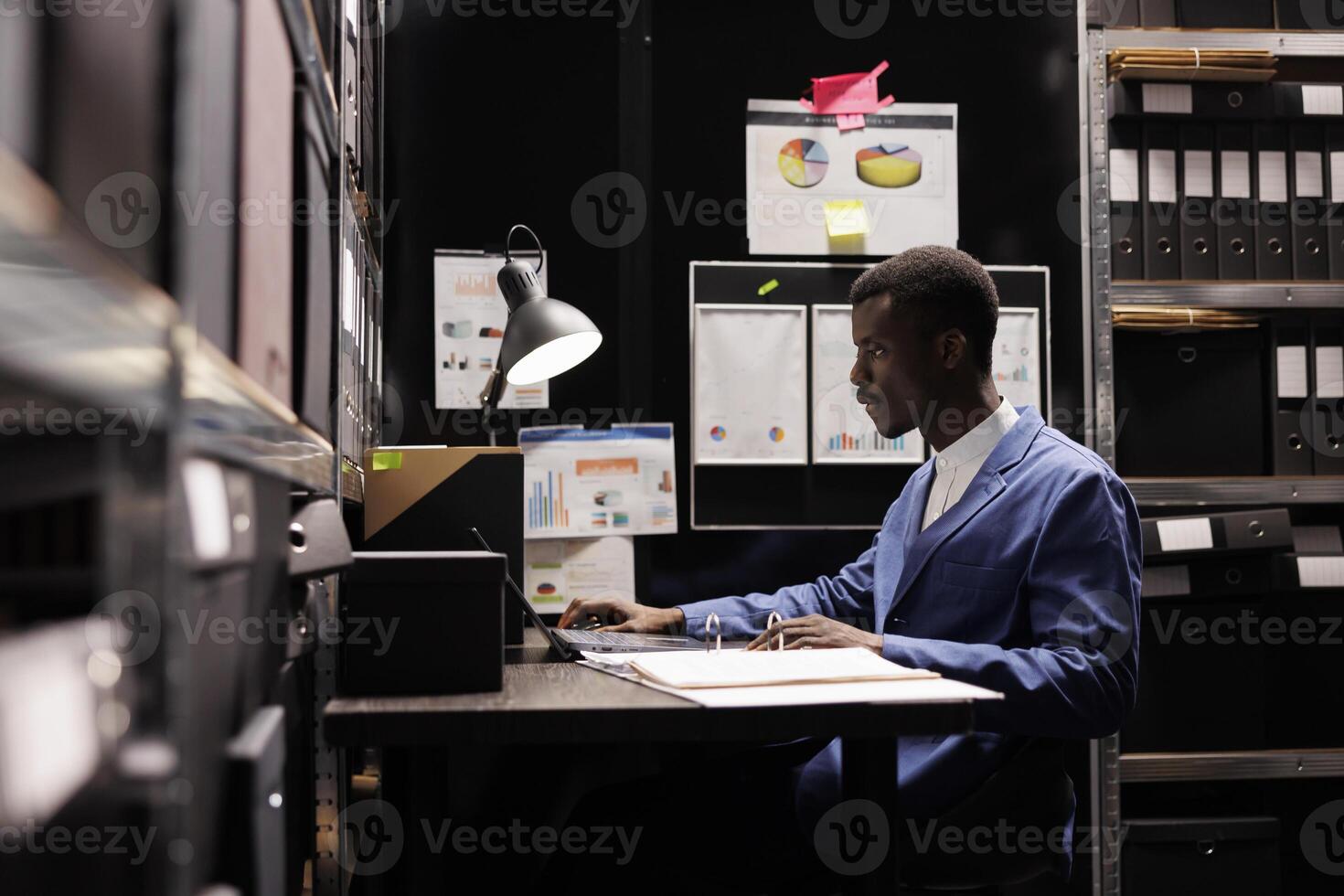 African american bookkeeper checking administrative files on laptop computer, working overtime in corporate depository. Businessman analyzing bureaucracy report in storage room photo