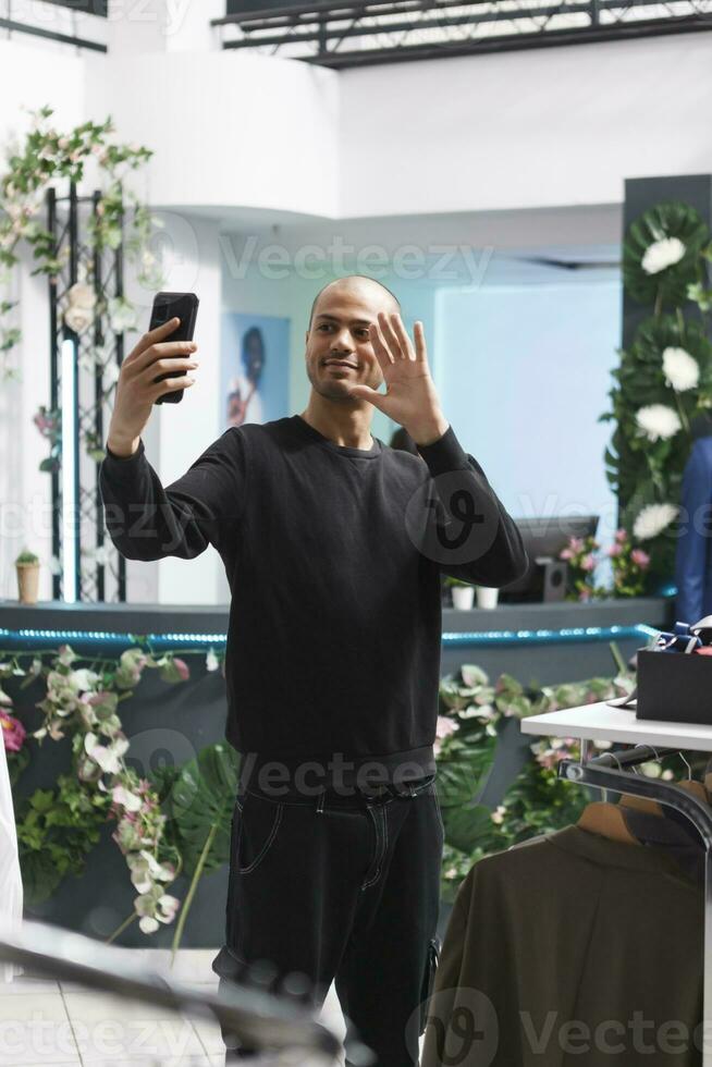 Shopping mall boutique arab man client waving hi while making selfie on smartphone. Young smiling blogger greeting social media followers while live broadcasting in clothing store photo