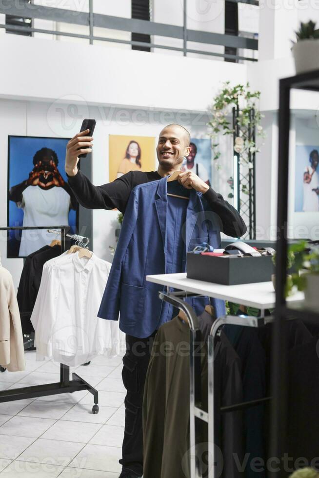 sonriente árabe Moda hombre de influencia utilizando teléfono inteligente frente cámara mientras demostración atuendo en percha y promoviendo vestir marca en boutique. ropa Tienda cliente tomando selfie en móvil teléfono foto
