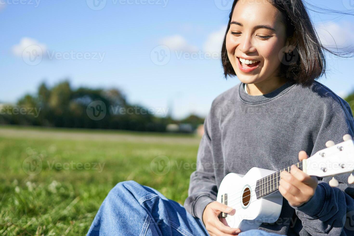 Happy people and hobbies. Smiling asian girl playing ukulele guitar and singing, sitting in park outdoors on blanket photo