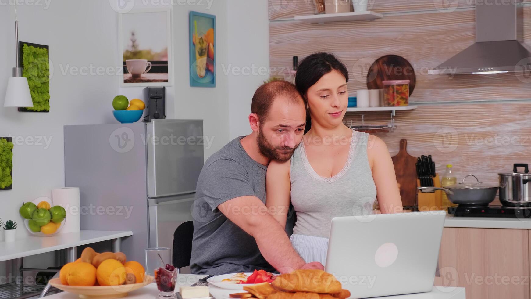 Happy couple surfing on internet using laptop during breakfast in kitchen. Married husband and wife in pajamas using internet web online modern technology, smiling and happy in the morning. Reading news browsing internet photo