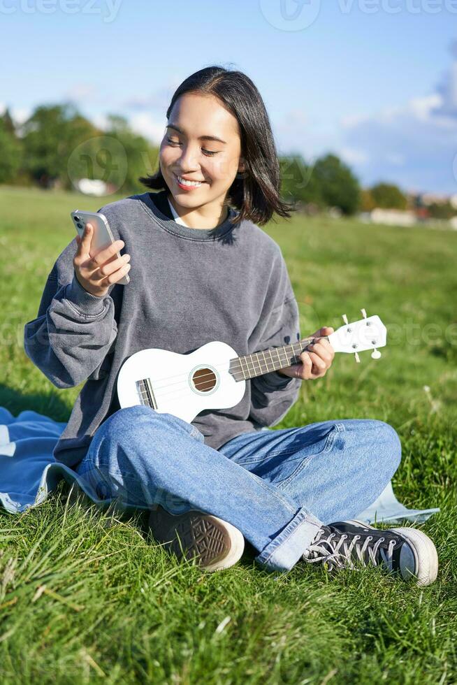 Vertical shot of smiling asian girl with smartphone, playing ukulele, reading chords or lyrics while singing, relaxing outdoors. Lifestyle and people concept photo