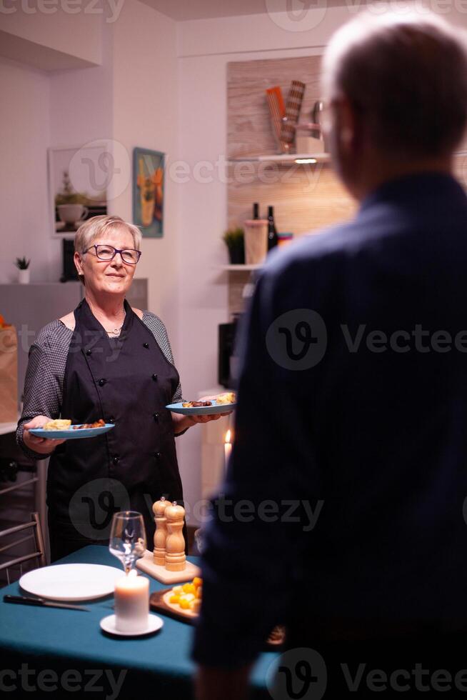 Senior couple having a conversation in kitchen. Elderly aged woman wearing apron. Elderly old couple talking, sitting at the table in kitchen, enjoying the meal, celebrating their anniversary photo